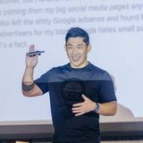 A smiling Asian man in a dark t-shirt holds a clicker while presenting in front of a screen with blurred text.