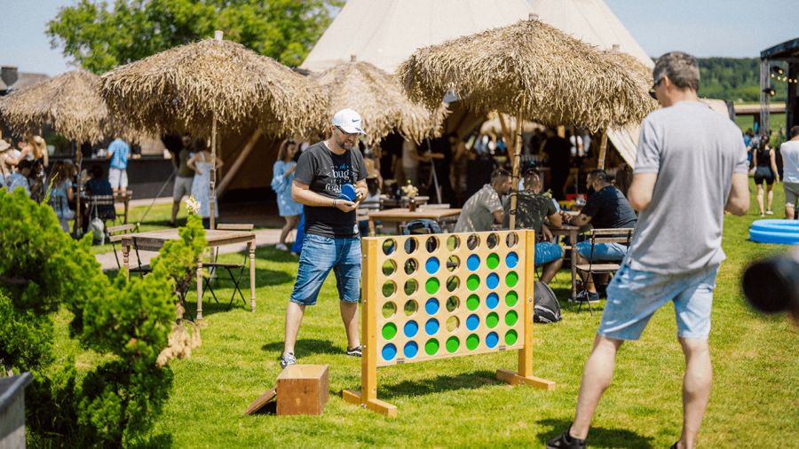 Two men play a giant Connect Four game on a sunny grassy lawn with people and tents in the background.