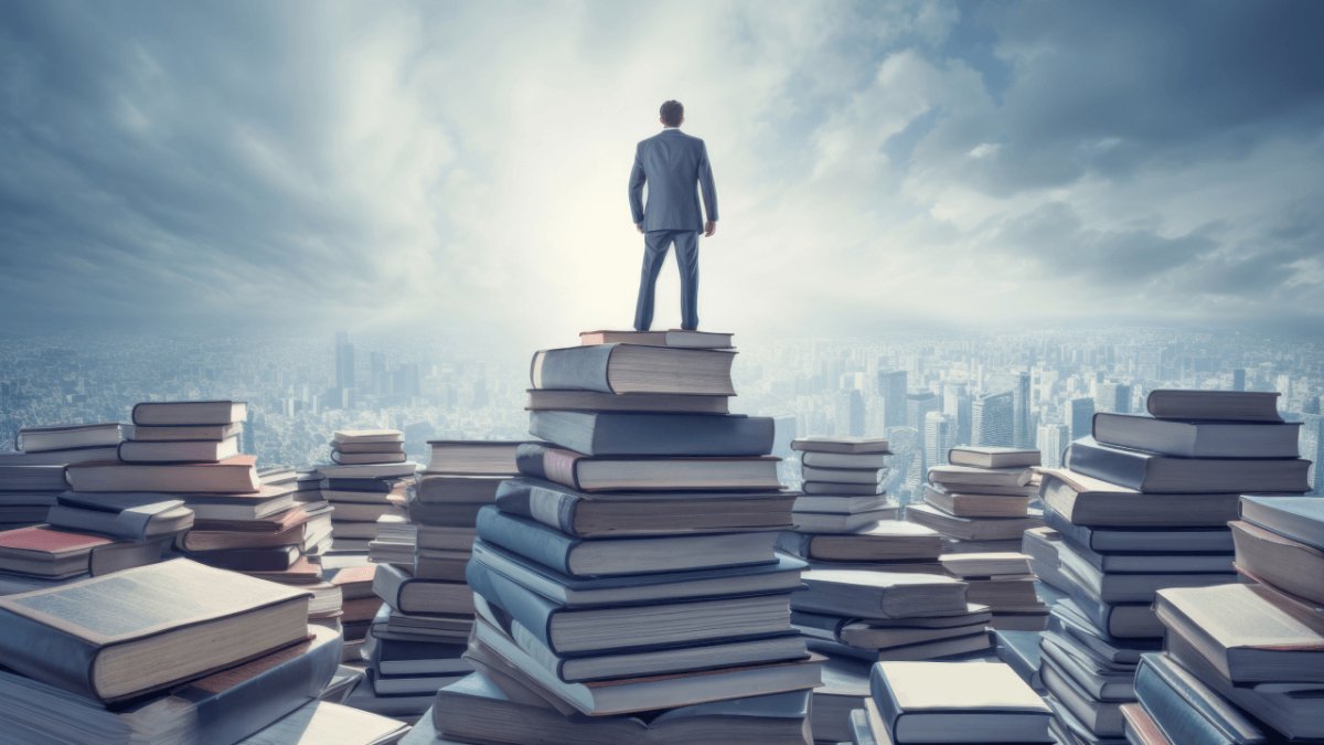 A man stands on a tall stack of books, looking out at a city skyline and a landscape of other book piles.