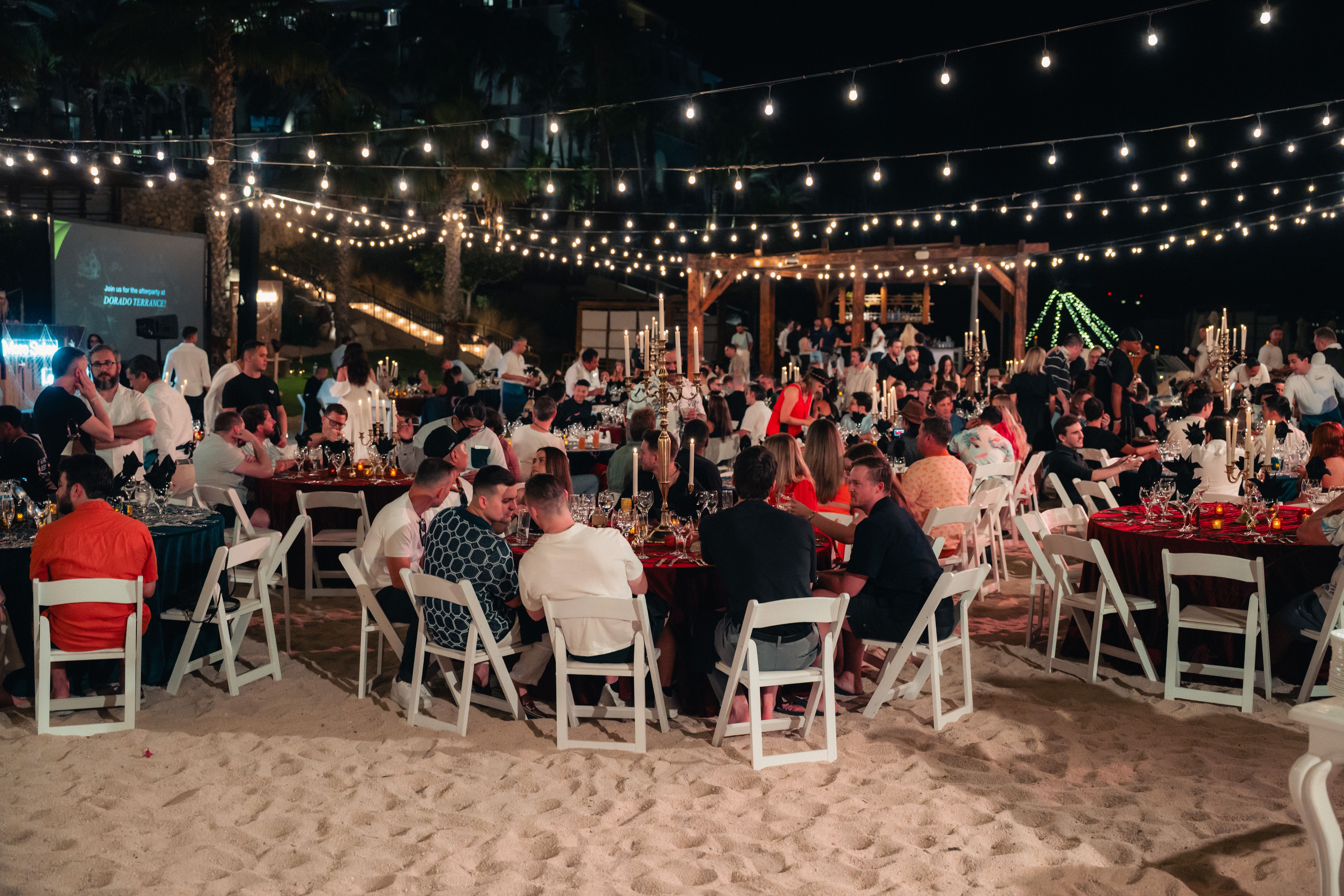 Guests dine at tables on a sandy beach at night, illuminated by string lights.