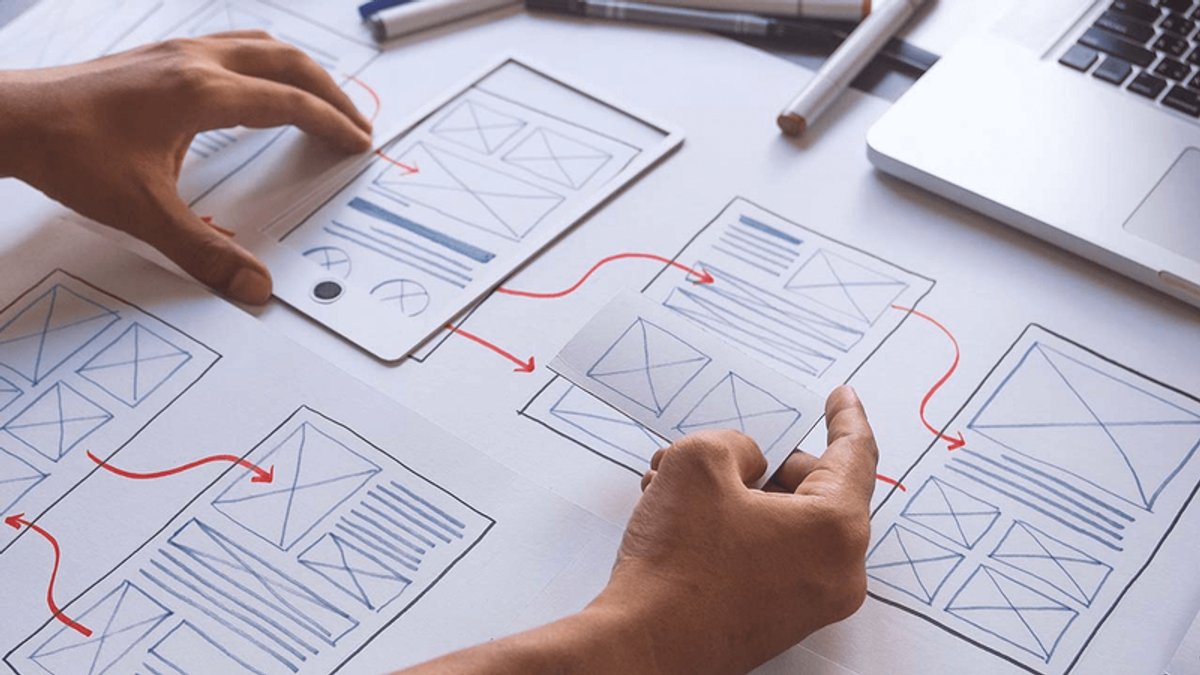Hands arranging paper wireframes for UI/UX design with red flow arrows.