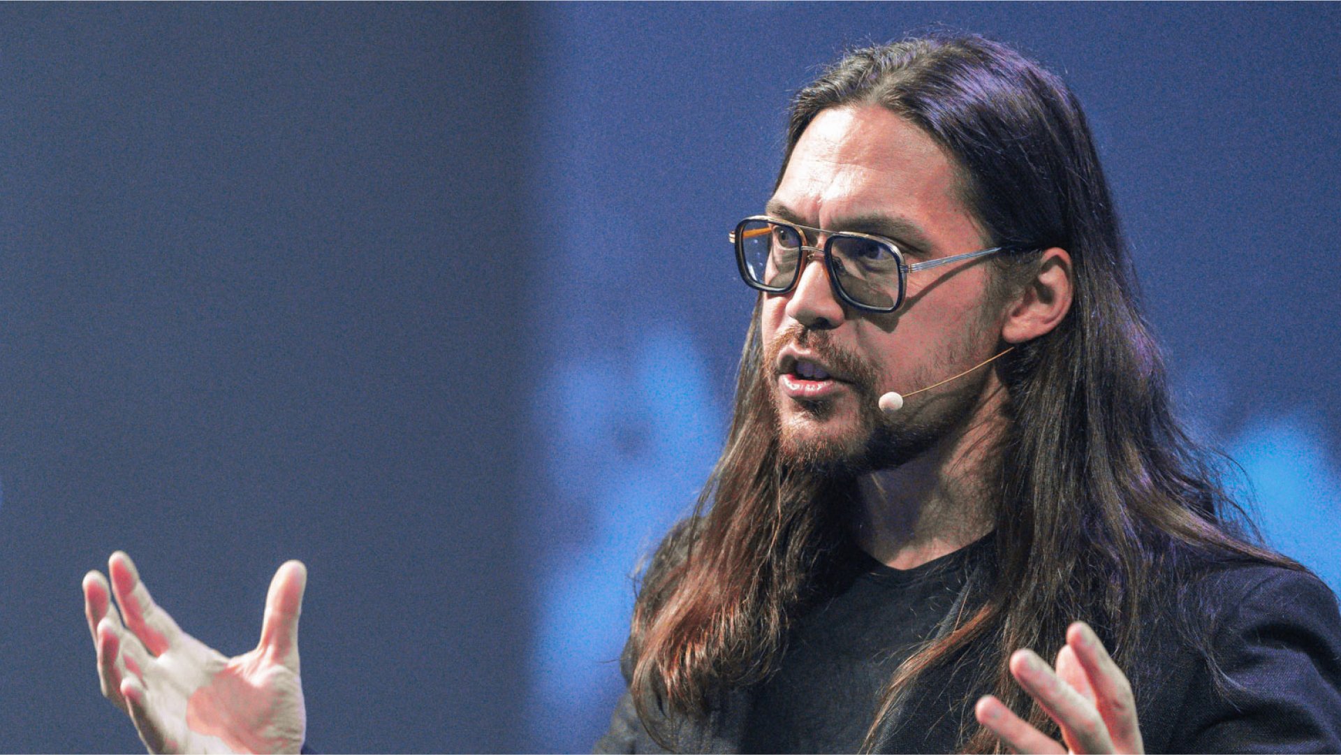A man with long dark hair, a beard, and glasses speaks into a microphone, gesturing with his hands.