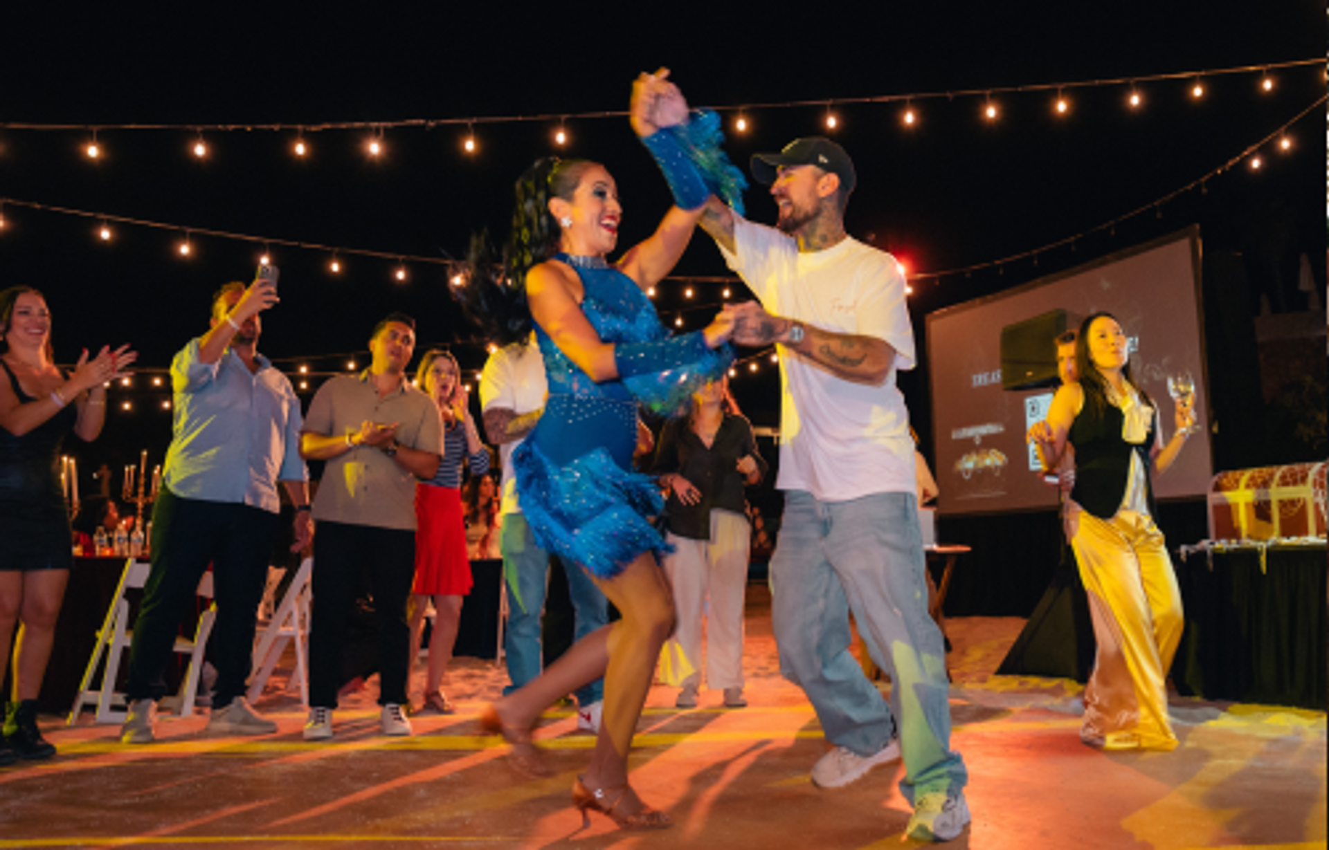 A man and woman dance energetically under string lights at an outdoor night event, watched by guests.
