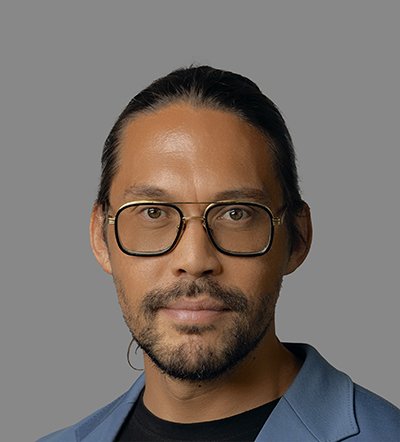 A man with pulled-back dark hair, a beard, and gold-framed glasses wears a blue jacket.