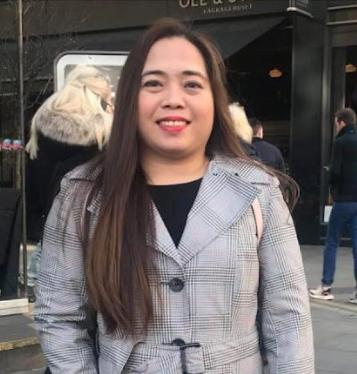 Mujer sonriente con cabello castaño largo vistiendo un abrigo gris de cuadros al aire libre.