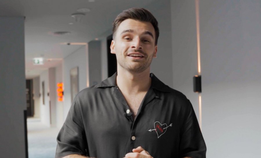 Un jeune homme souriant portant un t-shirt noir avec un motif cœur et flèche se tient dans un couloir moderne.