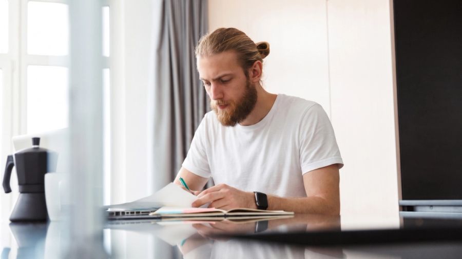 A bearded man in a white t-shirt writes in a notebook at a table, with a laptop and coffee pot nearby.