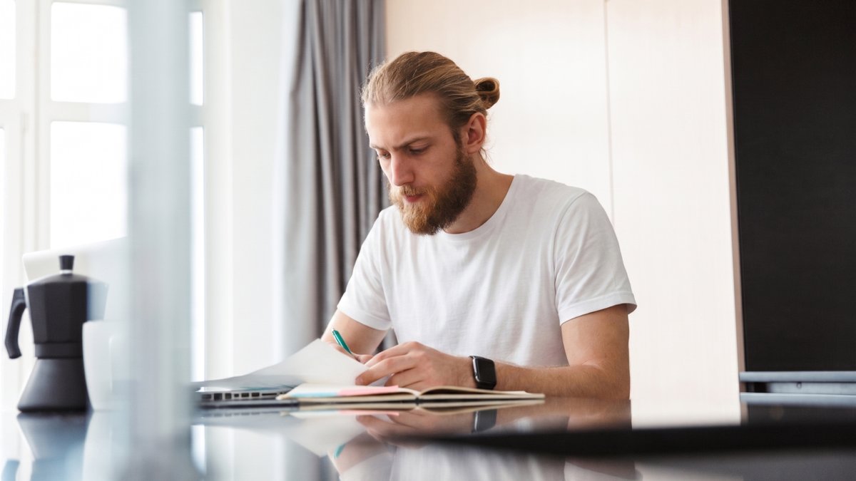 A bearded man in a white t-shirt writes in a notebook at a table, with a laptop and coffee pot nearby.