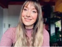 A smiling young woman with long blonde hair and bangs in a pink sweater.