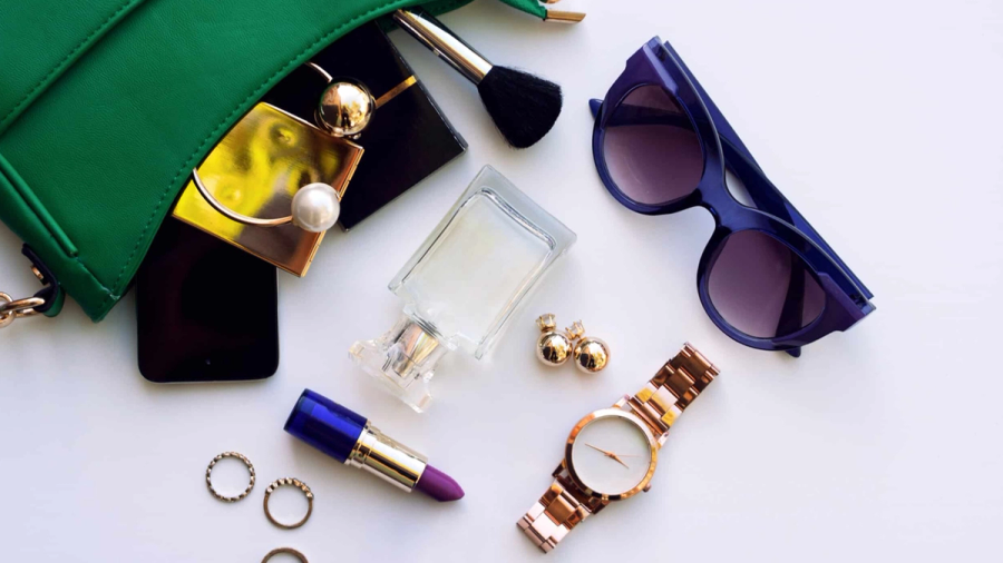 Green purse with various beauty and fashion accessories, including lipstick, perfume, sunglasses, a watch, and jewelry, on a white background.