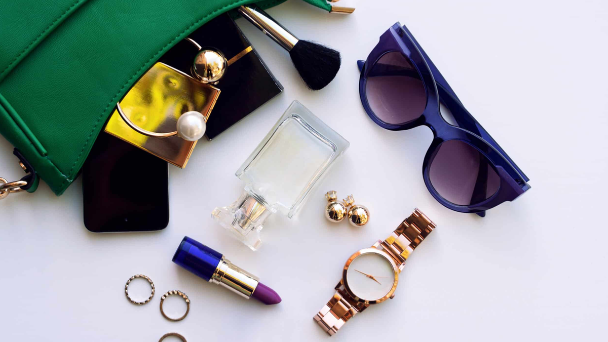 Green purse with various beauty and fashion accessories, including lipstick, perfume, sunglasses, a watch, and jewelry, on a white background.