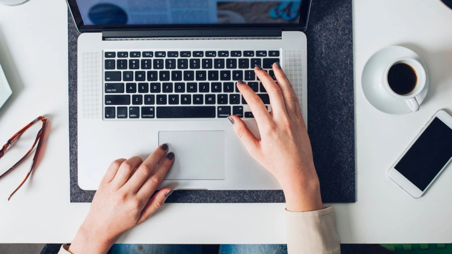Hands typing on a laptop, with a coffee cup, smartphone, and glasses on a white desk.