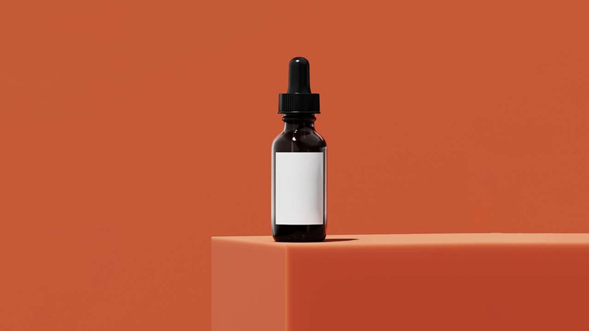 A dark brown dropper bottle with a blank white label stands on an orange block against an orange background.