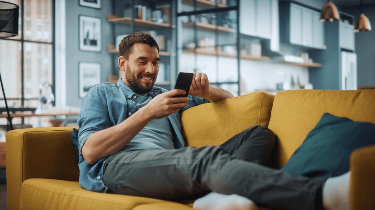 A smiling man relaxes on a yellow couch, looking at his phone.
