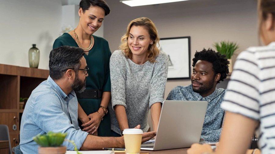 Four smiling professionals collaborate around a laptop in an office meeting.