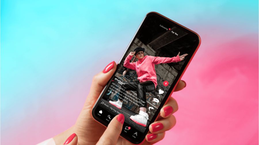 A hand with red nails holds a phone displaying a social media video of a dancer in a pink hoodie, against a blue and pink background.
