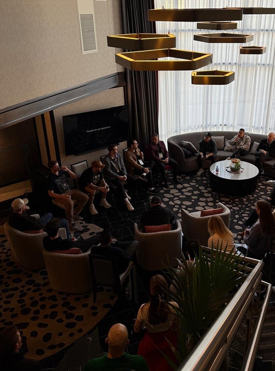 An overhead view of people gathered in a modern lounge for a discussion, with some seated on a curved sofa and others in armchairs facing a screen displaying 'Thanks to our sponsors'.