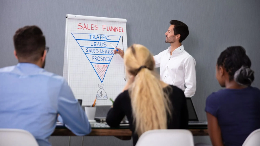 A man points to a sales funnel diagram on a flipchart, presenting to an audience.