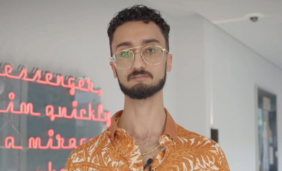 Homem com cabelo crespo escuro, barba e óculos veste camisa laranja estampada e corrente dourada, com letreiro neon rosa ao fundo.