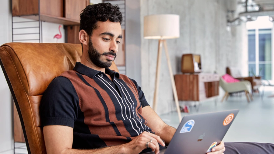 A bearded man works on a laptop while sitting in a brown leather chair.