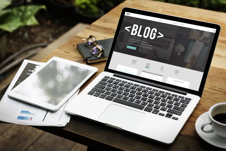 Laptop on a wooden table displaying a 'BLOG' webpage, with a tablet, papers, glasses, and coffee nearby.