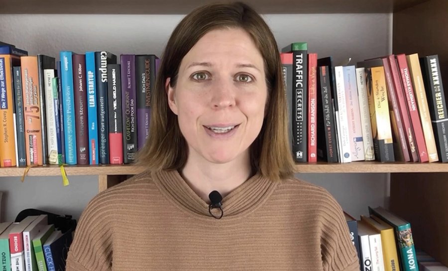 A smiling woman in a brown ribbed sweater with a lapel mic, standing in front of a bookshelf filled with books.