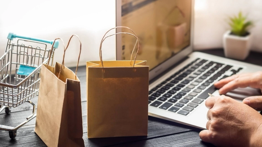 Online shopping scene with a person typing on a laptop, a miniature shopping cart, and brown paper bags.