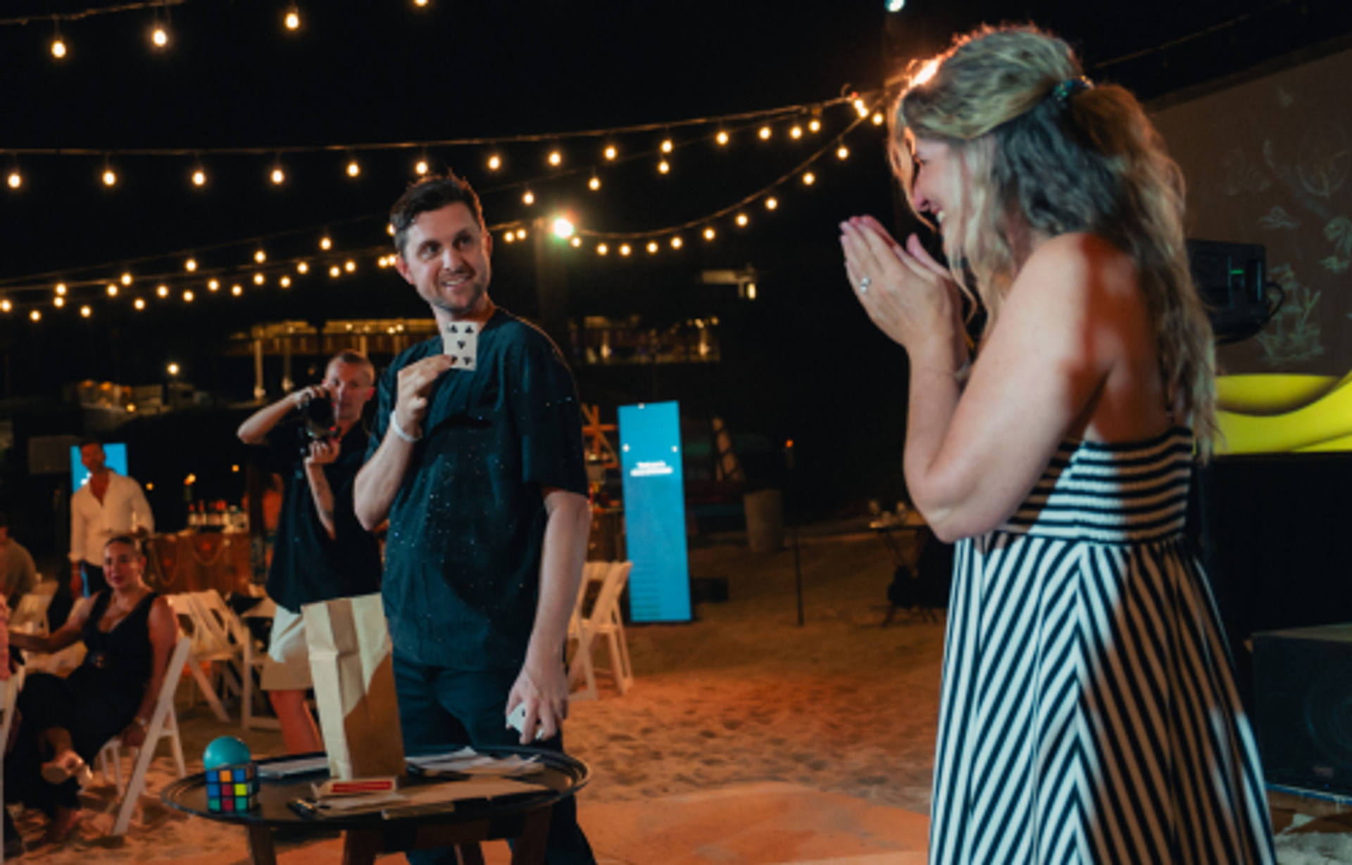 A magician holds up a card for a woman who claps and laughs in delight at an outdoor night event with string lights.