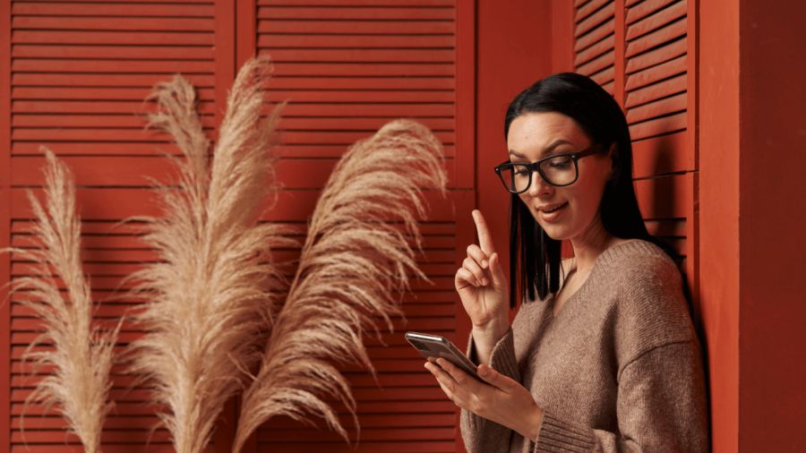 A woman in glasses looks at her phone, raising her finger as if having an idea, against a red wall with pampas grass.