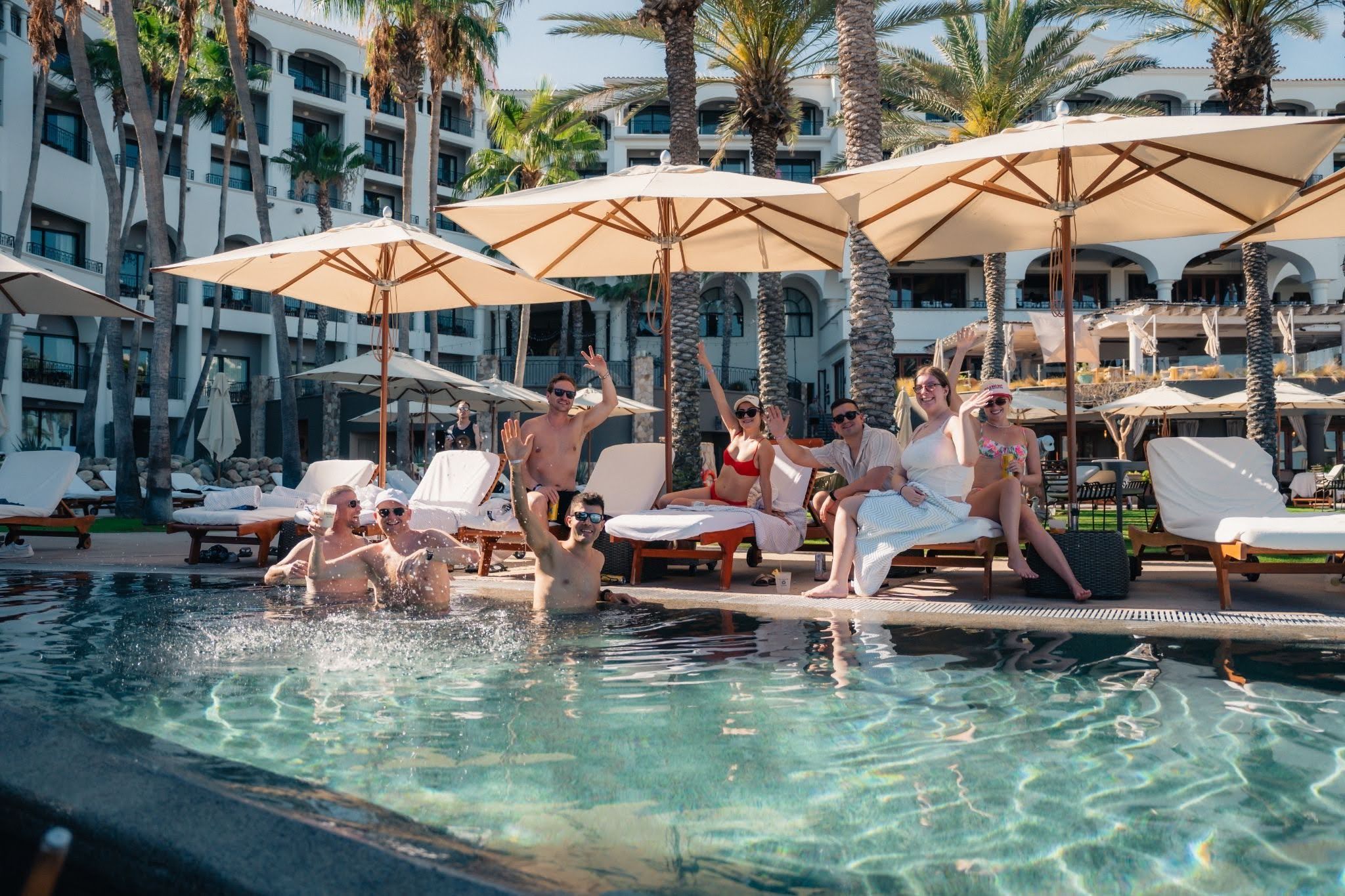 A group of friends smiling and waving while enjoying a sunny day at a resort pool, with some in the water and others on lounge chairs.