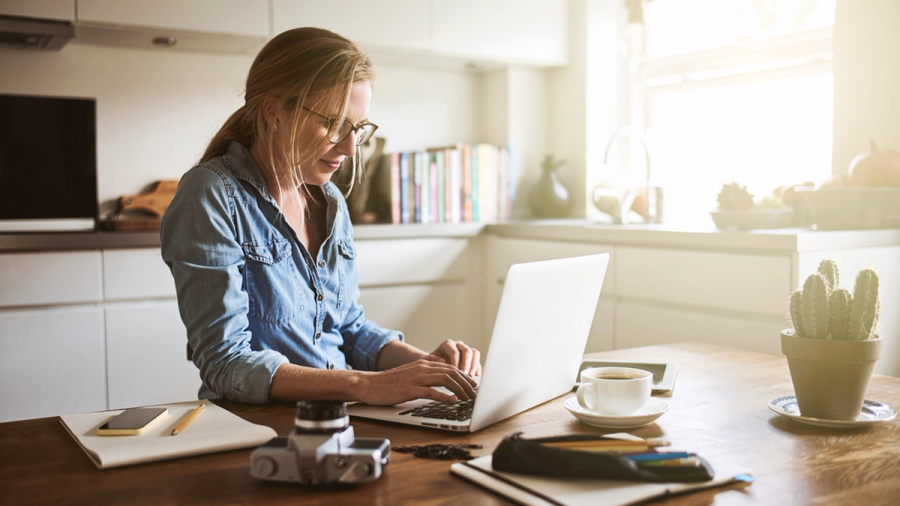 A woman in glasses types on a laptop at a bright kitchen table with coffee and a cactus.