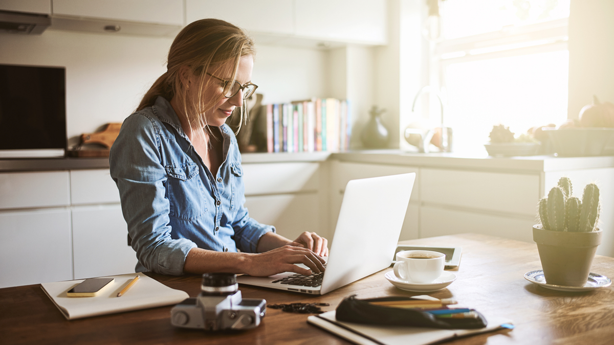 A woman in glasses types on a laptop at a bright kitchen table with coffee and a cactus.