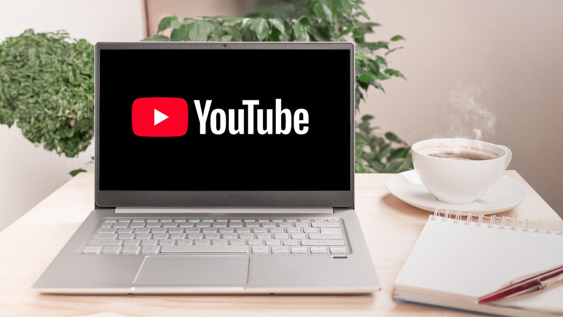 A laptop displaying the YouTube logo next to a steaming cup of coffee, a notebook, and a pen on a desk.