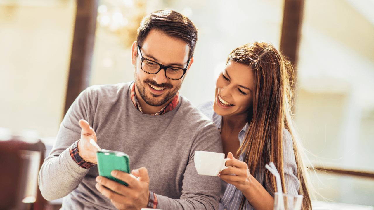 A smiling man shows a smartphone to a laughing woman holding a coffee cup.