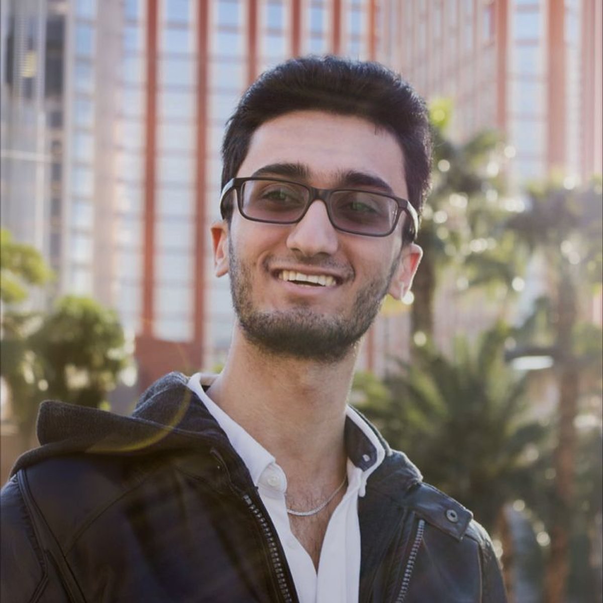 Smiling young man wearing glasses and a leather jacket, with blurred buildings and palm trees in the background.