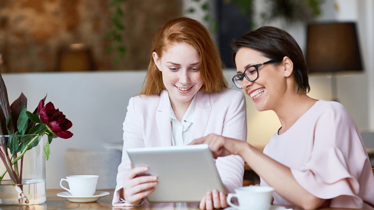 Two smiling women looking at a tablet together, one pointing at the screen.
