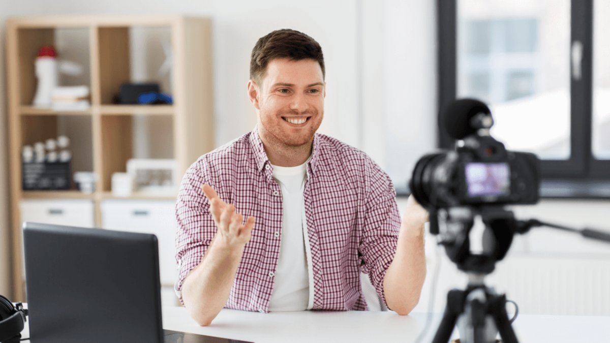 A smiling man records a video in a home studio with a camera on a tripod and a laptop.