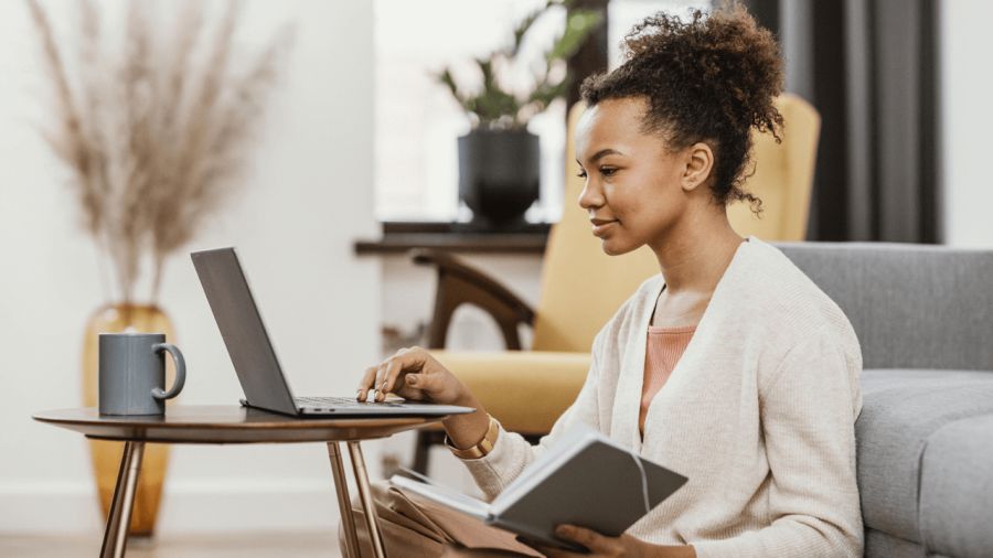 A young Black woman sits on the floor, typing on a laptop and holding a notebook.