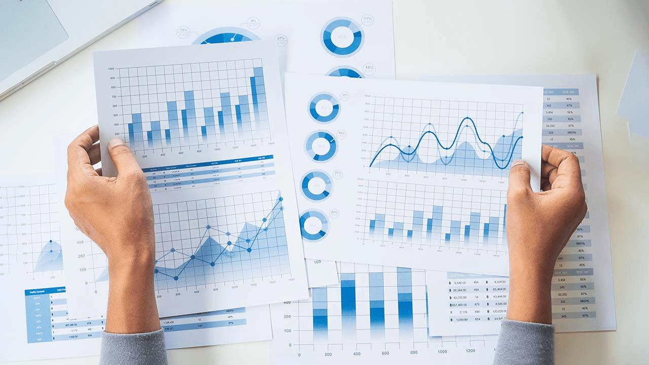 Hands holding two sheets of paper with blue charts and graphs, surrounded by more data reports.