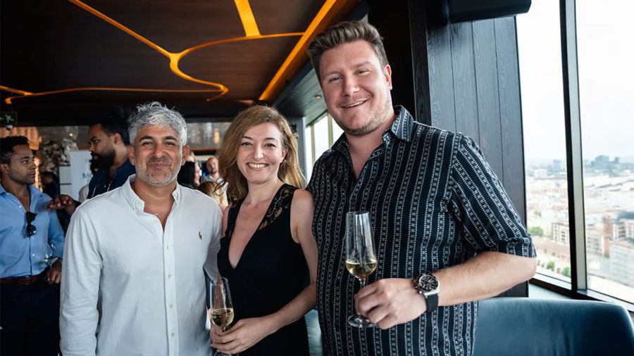 Three people smiling and holding drinks at a bar overlooking a city.