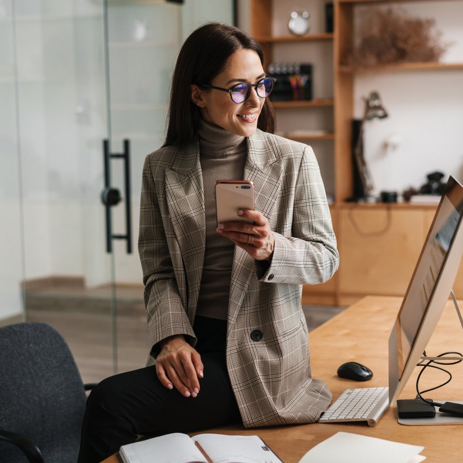 Smiling woman in a plaid blazer holds a smartphone on an office desk.