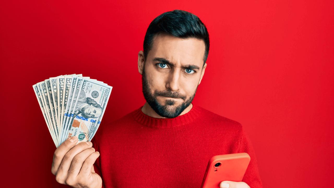 A frowning man in a red sweater holds a fan of dollar bills and a red smartphone against a red background.