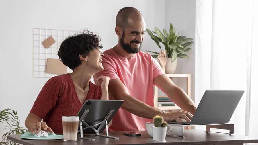 A laughing man and woman work side-by-side at a desk with a laptop and tablet.