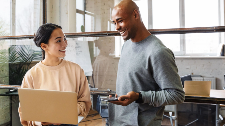 A smiling man and woman looking at each other in an office, holding a laptop and phone.