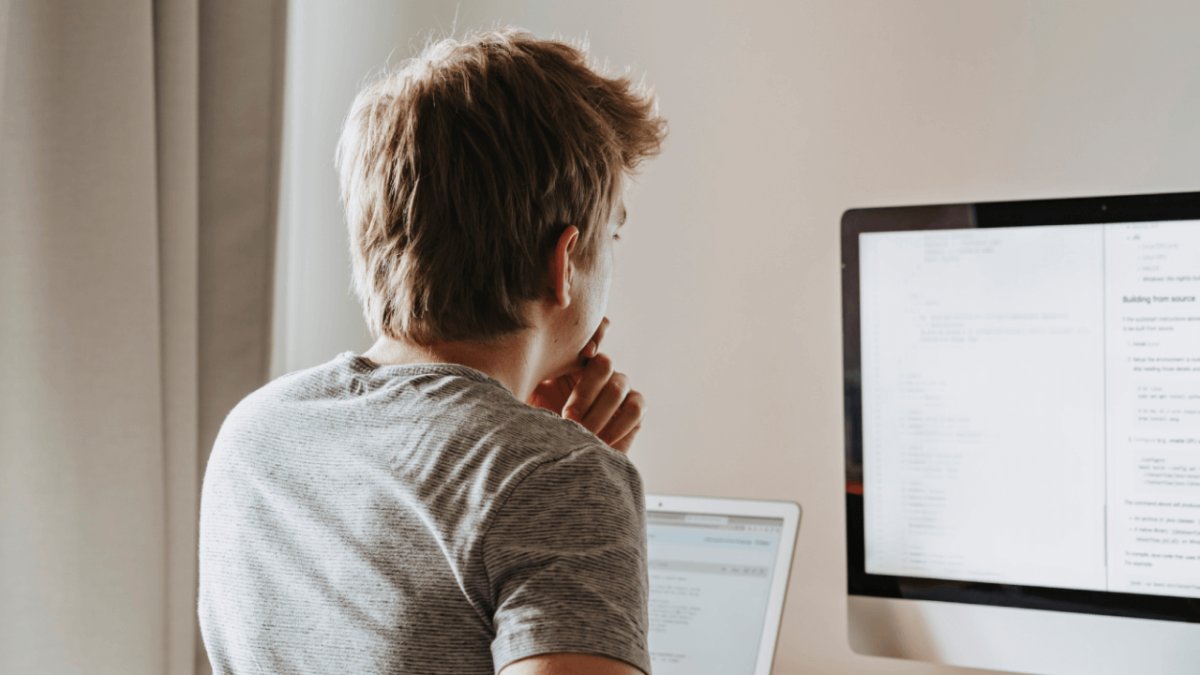 A person from behind works on code displayed on a monitor and laptop.