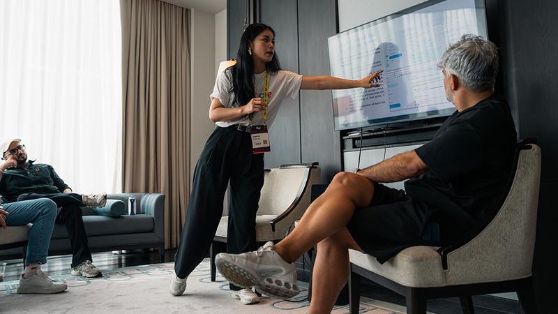 A woman points to a screen displaying data for a seated man, with two other people in the background.