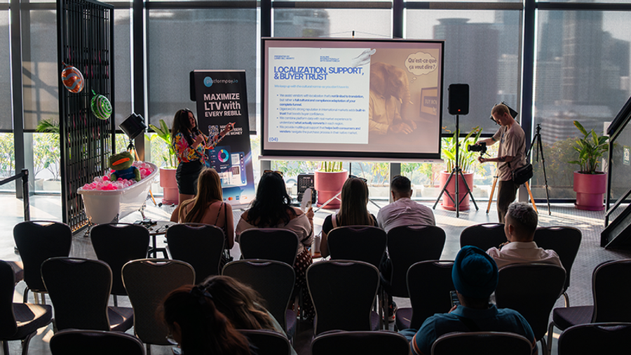 A woman gives a presentation to an audience in a sunlit room, with a large screen displaying slides and a videographer filming.