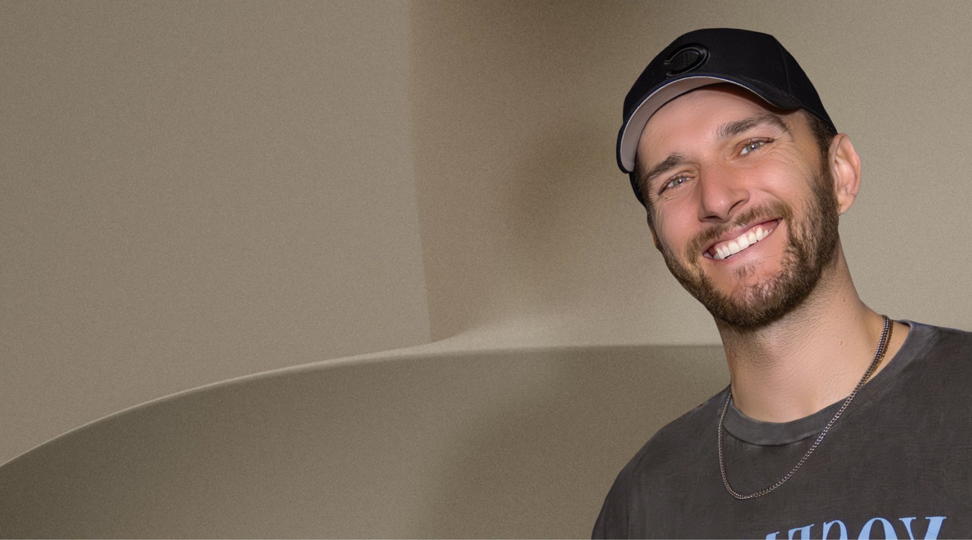 Un homme souriant portant une casquette noire et un t-shirt, avec une barbe et un collier.