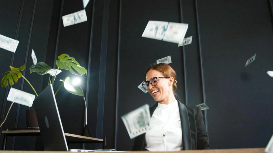 A smiling woman in glasses sitting at a desk with money falling around her.