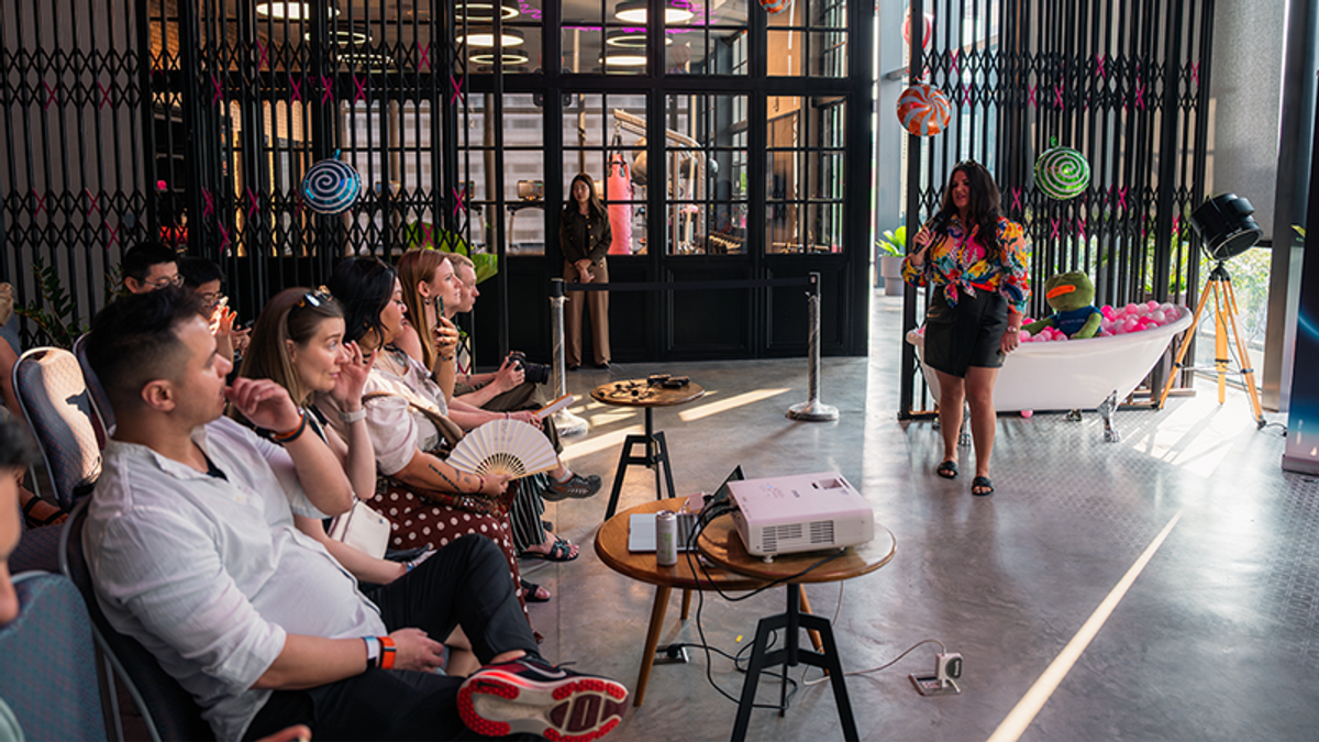 A woman speaks to an audience in a modern event space featuring a decorative bathtub and large lollipop props.